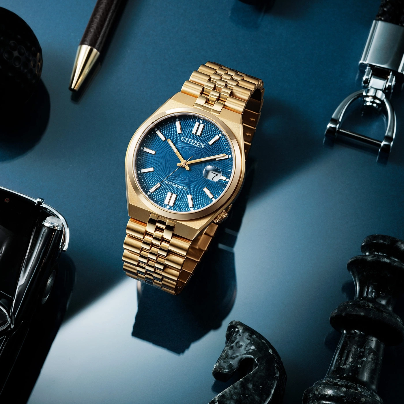 Goldene Citizen-Uhr mit blauem Zifferblatt, Stift, Schachfiguren und Schlüsselanhänger auf blauer Oberfläche.