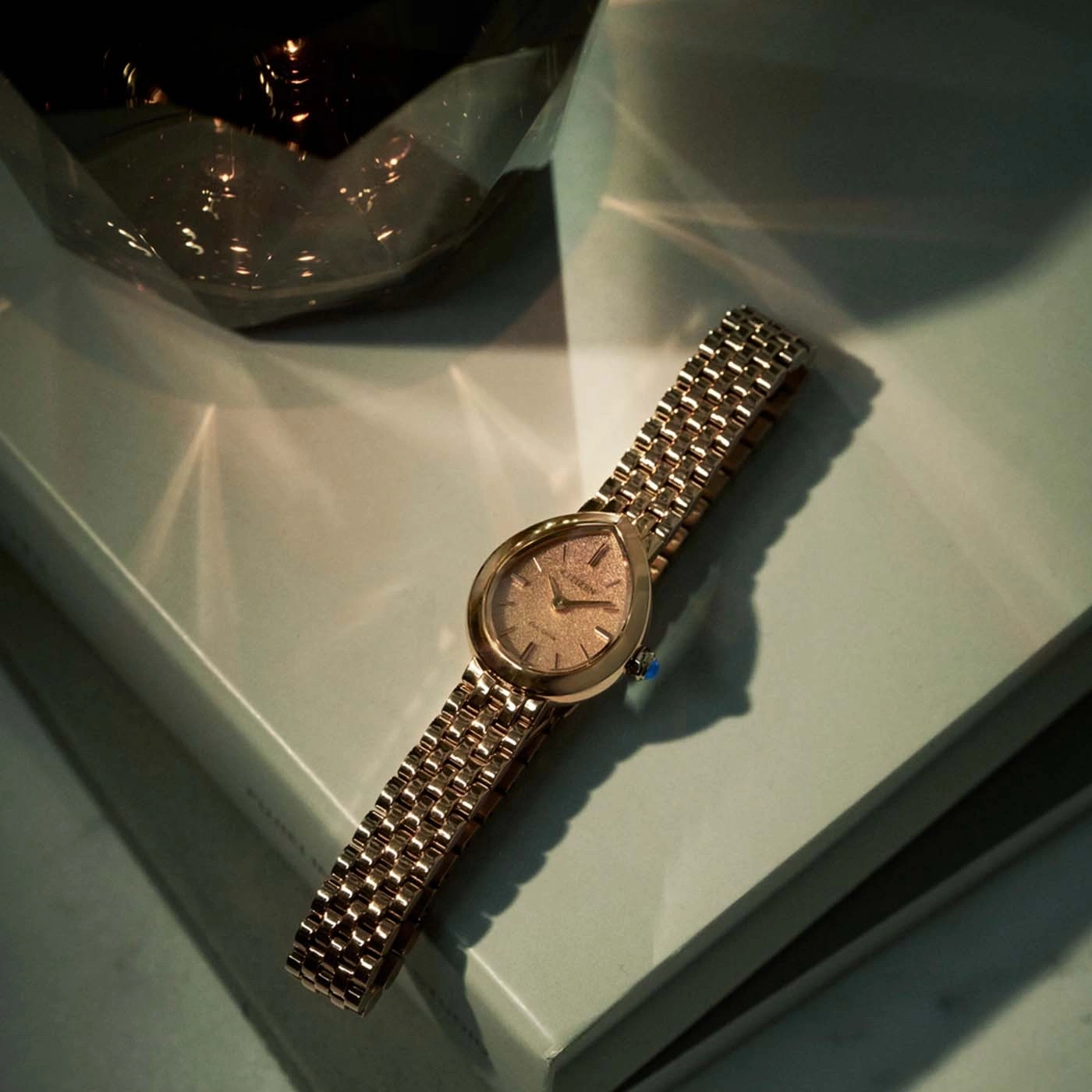 Eine goldene Uhr mit gemustertem Zifferblatt liegt auf einer hellgrünen Oberfläche.