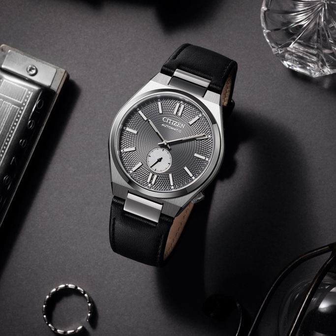 A stylish Citizen automatic watch with a black leather strap rests on a dark surface, surrounded by a harmonica, a crystal glass, a ring, and eyeglasses.
