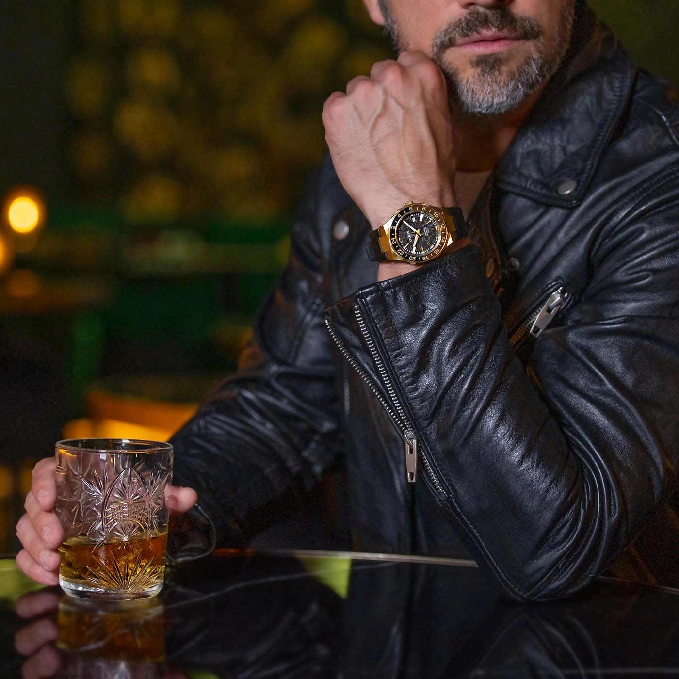 Mann in Lederjacke mit Uhr hält Whiskeyglas.