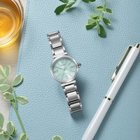 Eine mintgrüne Citizen Eco-Drive Uhr mit silbernem Band liegt auf einer blauen strukturierten Oberfläche neben einem weißen Stift und einem Glas mit bernsteinfarbener Flüssigkeit.