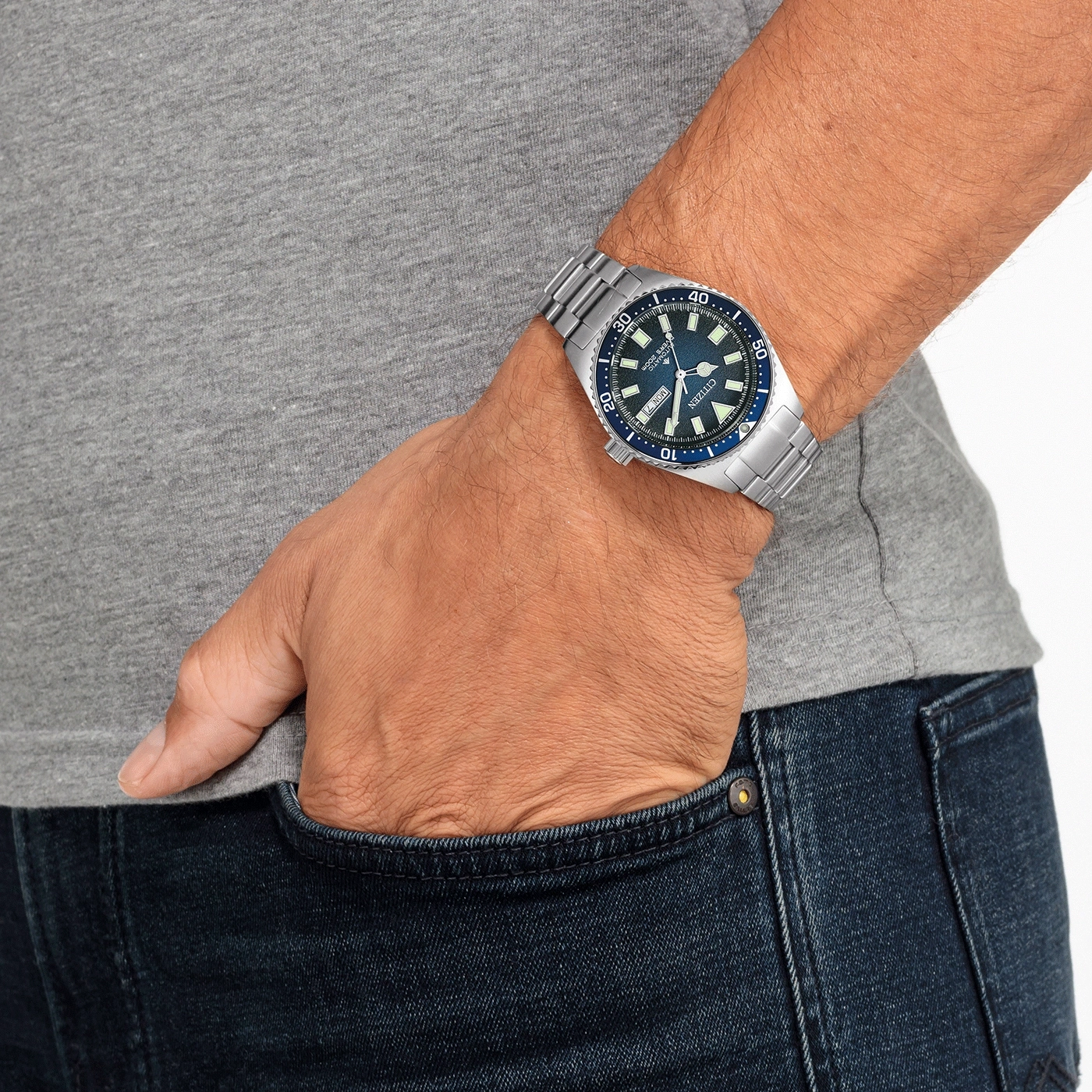 Mann mit Uhr, Hand in der Tasche, blaue Jeans und graues Hemd.