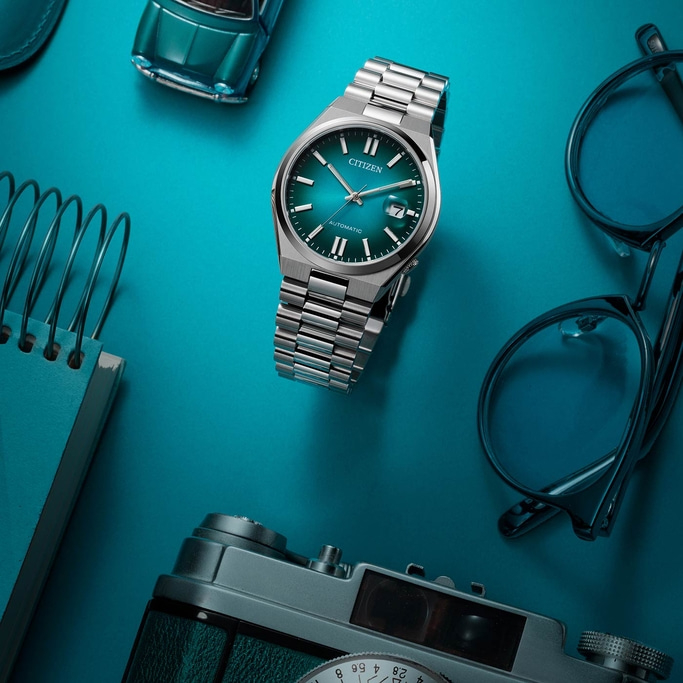 Citizen-Uhr, Brille, Kamera, Spielzeugauto und Notizbuch auf blauem Untergrund.