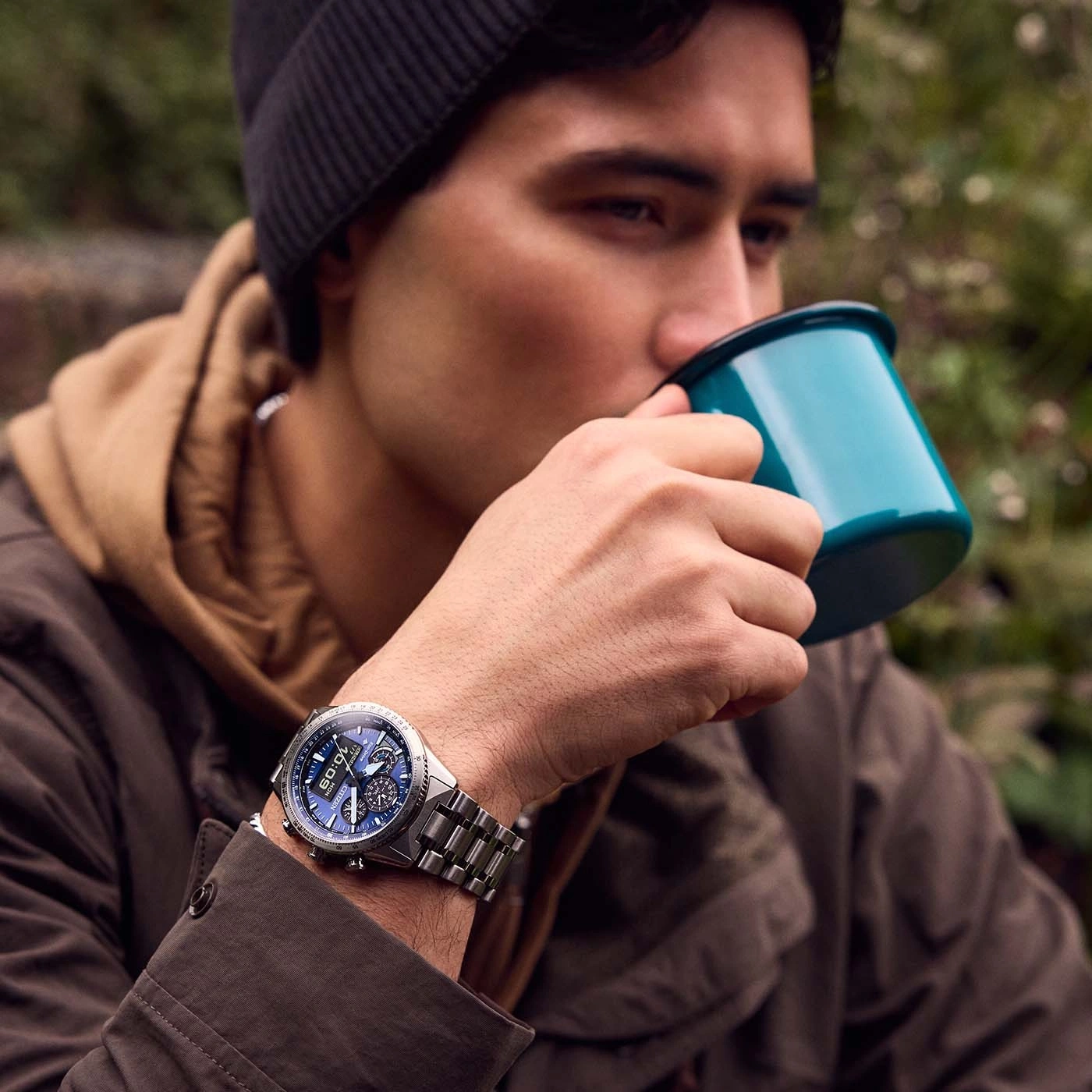 Mann trinkt aus einer Tasse, trägt eine Uhr und eine Mütze in der Natur.