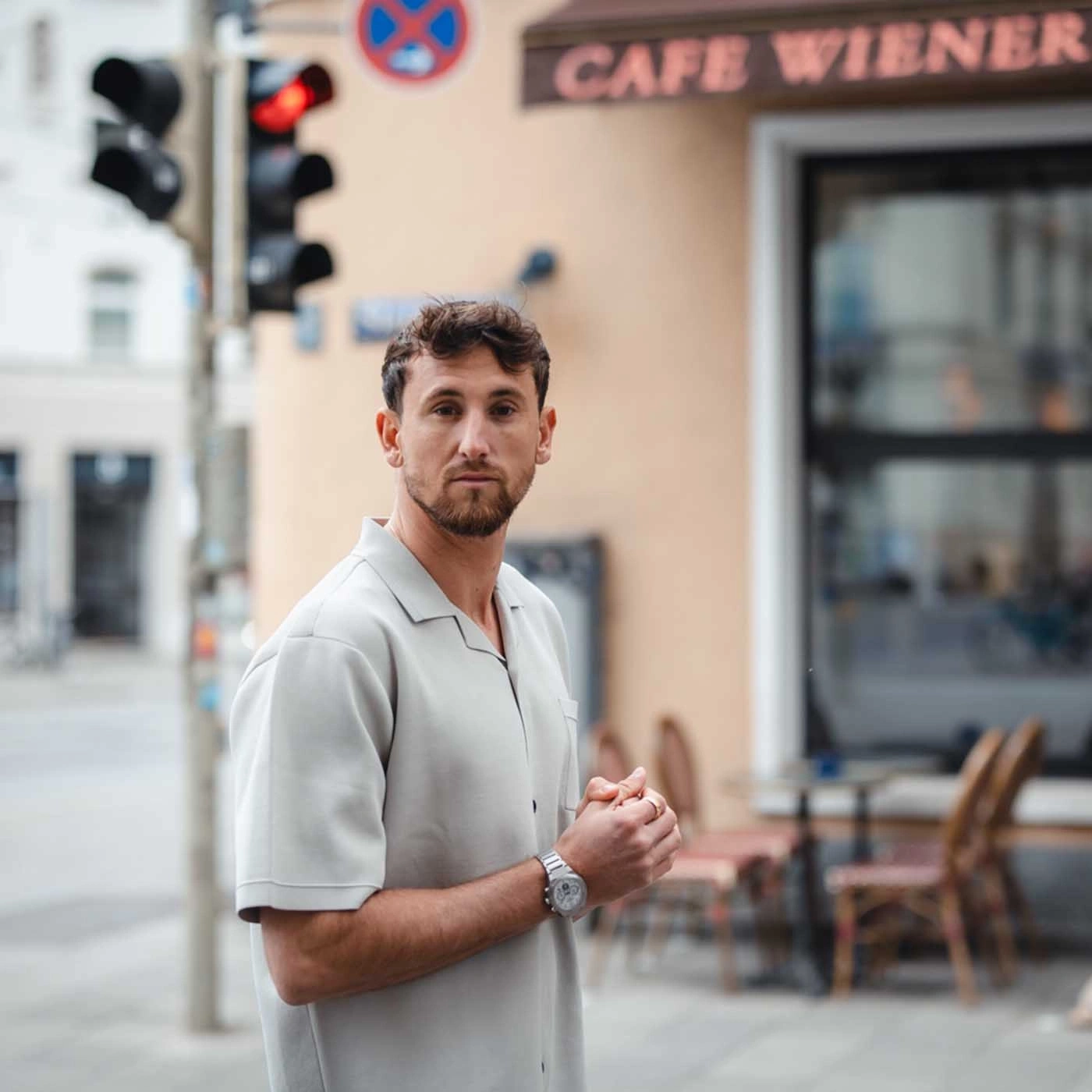 Mann vor einem Café, trägt ein helles Hemd und eine Uhr, schaut in die Kamera.