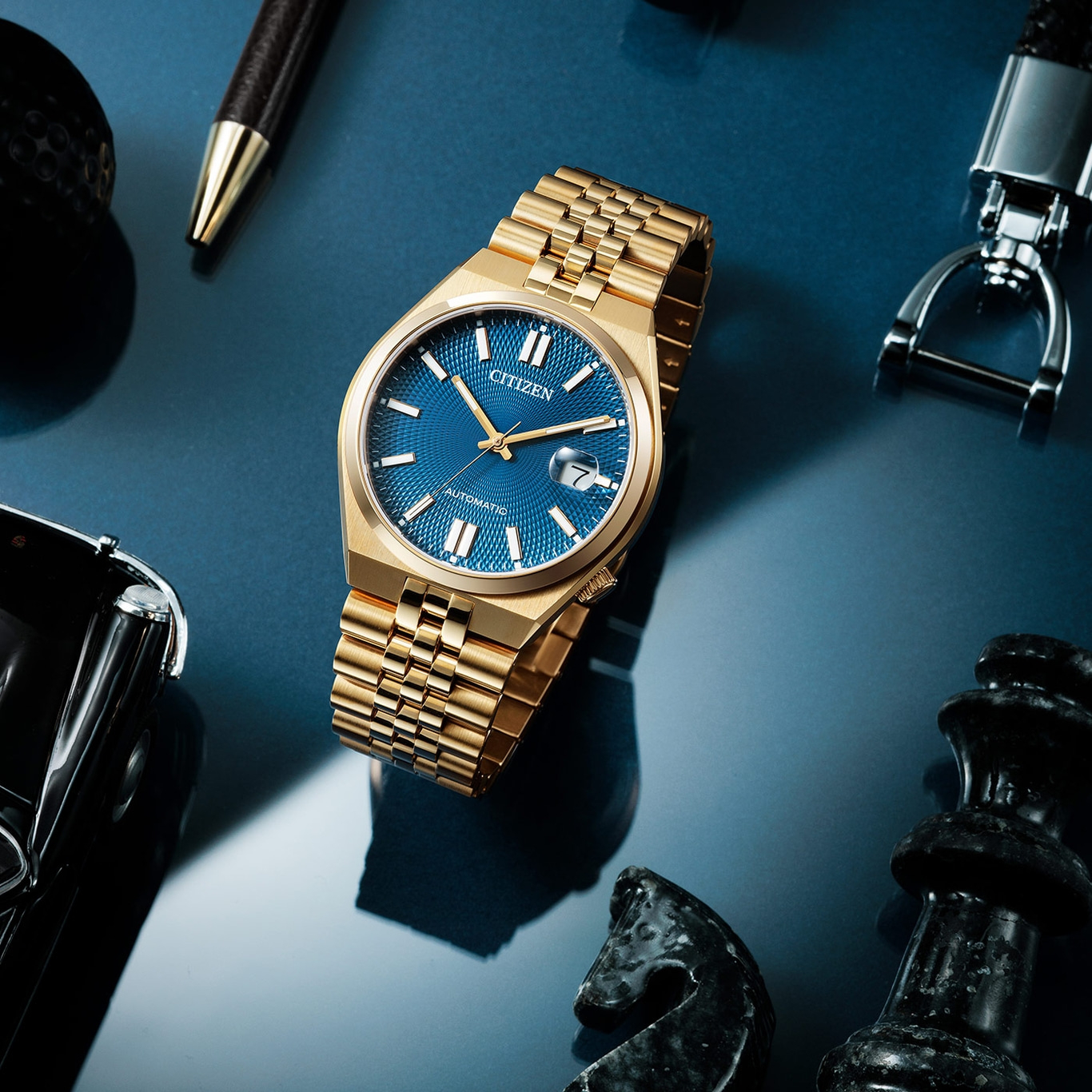 Goldene Citizen-Uhr mit blauem Zifferblatt, Stift, Schachfiguren und Schlüsselanhänger auf blauer Oberfläche.
