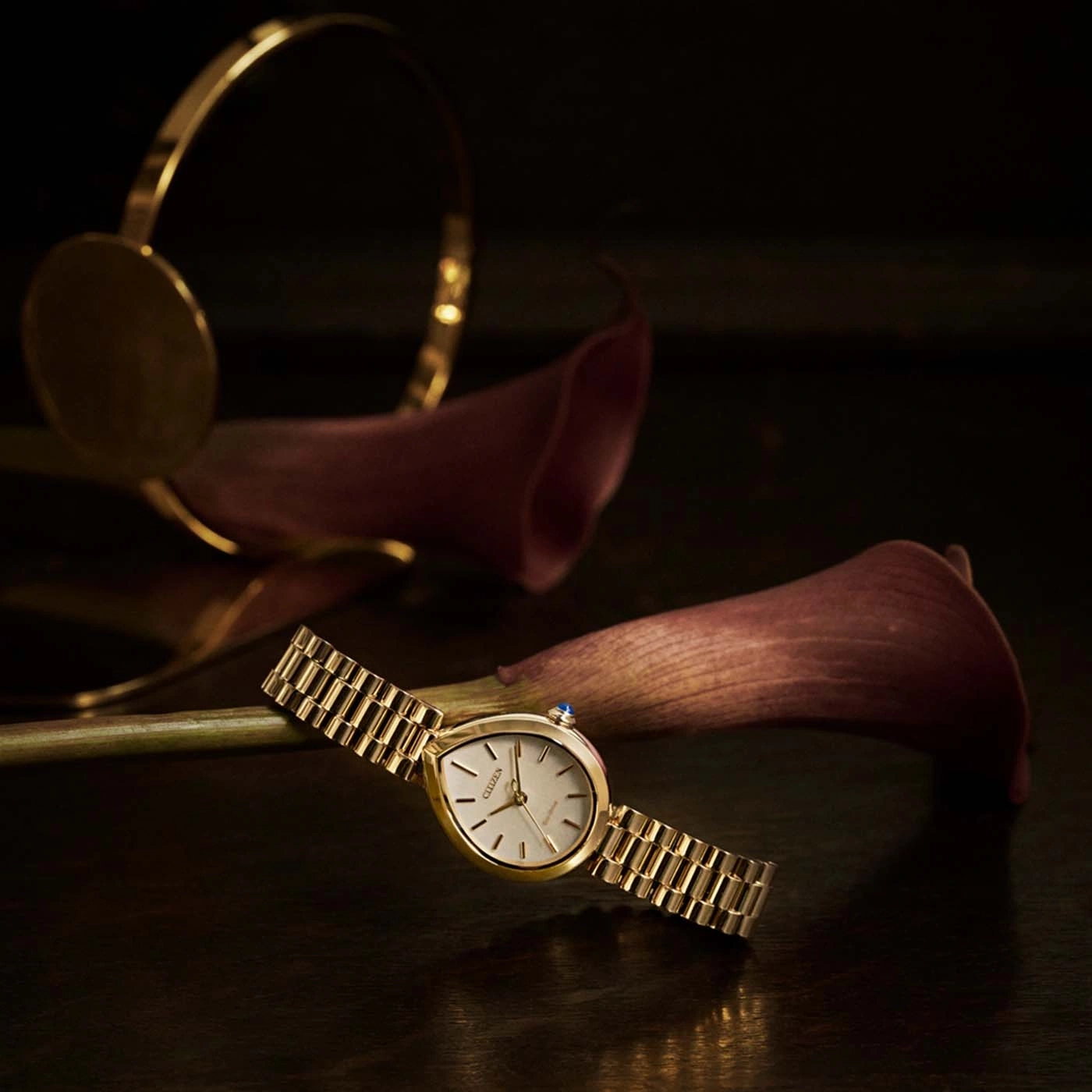 Eine goldene Uhr mit Armband liegt auf dunkler Oberfläche mit Blumen.