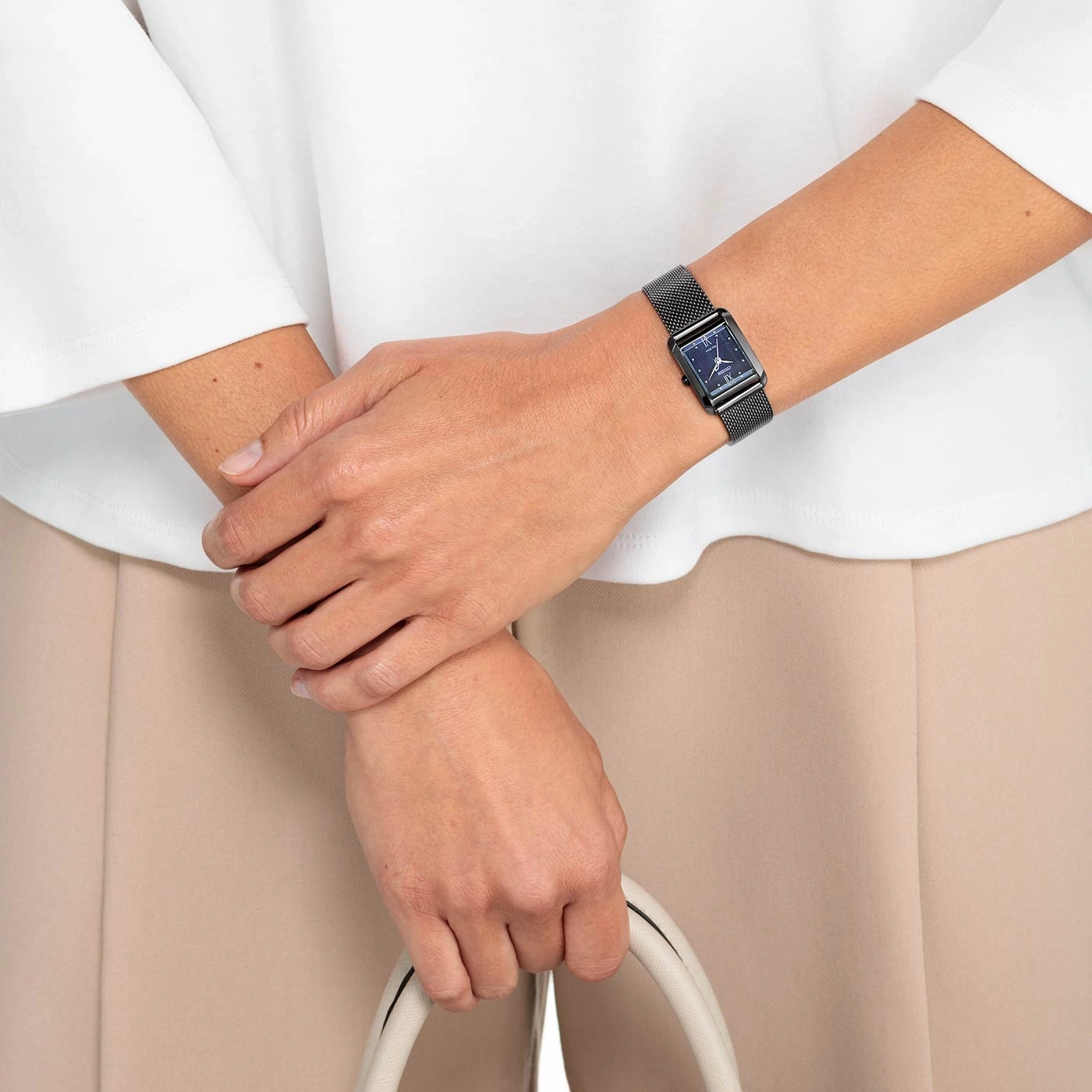 Frau mit schwarzer Uhr, hält weiße Tasche, beige Hose, weißes Hemd.