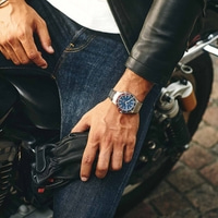 Mann mit Uhr, Lederjacke und Jeans, hält Handschuhe auf einem Motorrad.
