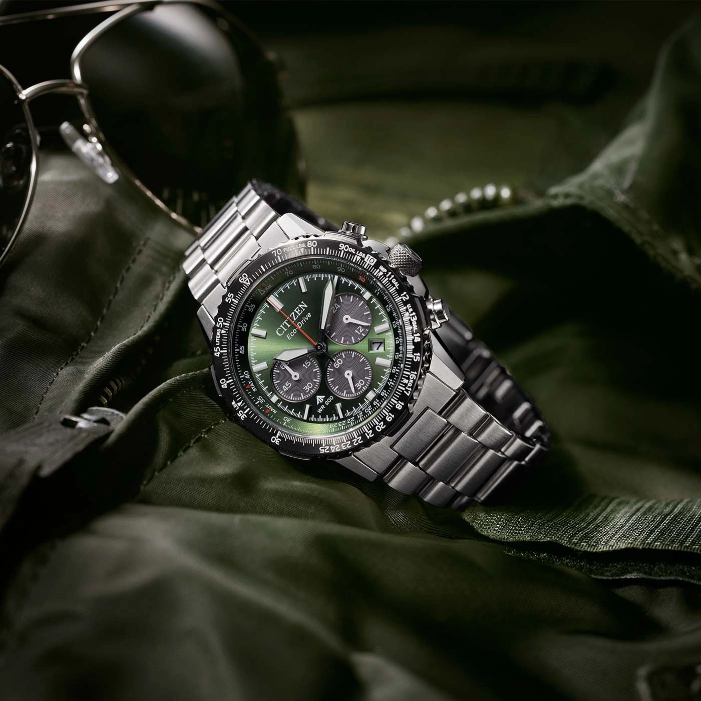 Citizen-Uhr mit grünem Zifferblatt und Metallband auf grünem Stoff mit Sonnenbrille.