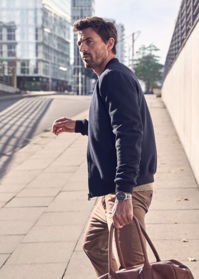 Mann geht auf dem Bürgersteig, hält eine Tasche, trägt eine Jacke und eine Uhr.