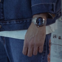 Mann trägt blaue Jeansjacke, weißes T-Shirt und Jeans mit Uhr