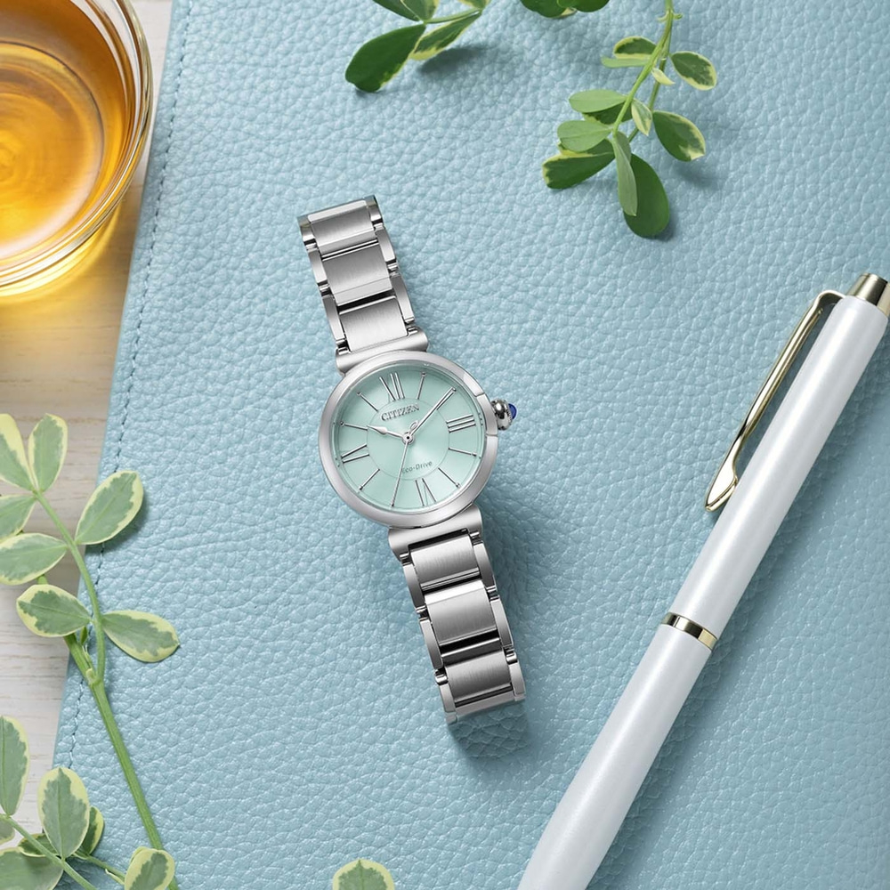 Eine mintgrüne Citizen Eco-Drive Uhr mit silbernem Band liegt auf einer blauen strukturierten Oberfläche neben einem weißen Stift und einem Glas mit bernsteinfarbener Flüssigkeit.