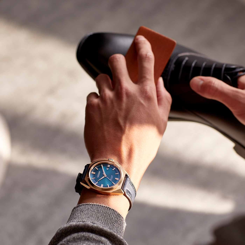 Mann poliert schwarzen Schuh, trägt goldene Uhr mit blauem Zifferblatt.