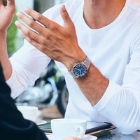 Mann mit Uhr, gestikuliert mit Händen, sitzt an einem Tisch mit Kaffeetassen.