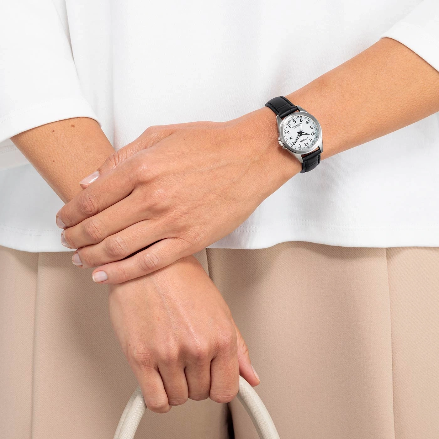 Frau mit Uhr, hält eine Tasche, mit weißem Hemd und beiger Hose.