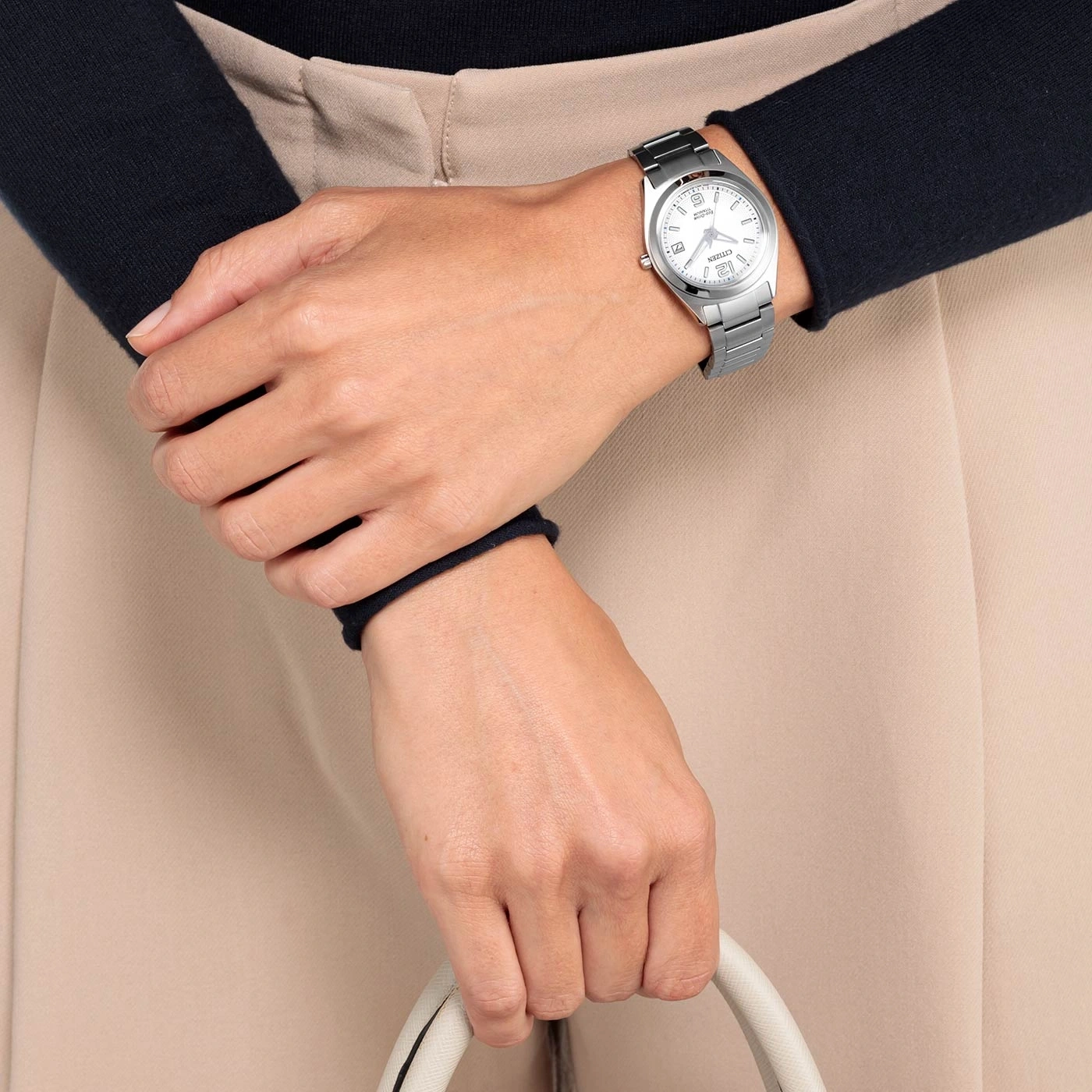 Frau mit silberner Uhr, hält eine weiße Handtasche, beige Hose.
