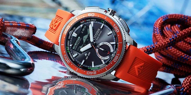 Eine sportliche Citizen Eco-Drive Uhr mit orangefarbenem Band liegt neben nautischen Seilen und Beschlägen auf einer reflektierenden Oberfläche.