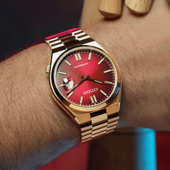 A close-up shot shows a gold Citizen watch with a striking red face on a hairy wrist, accented by moody red and blue lighting.
