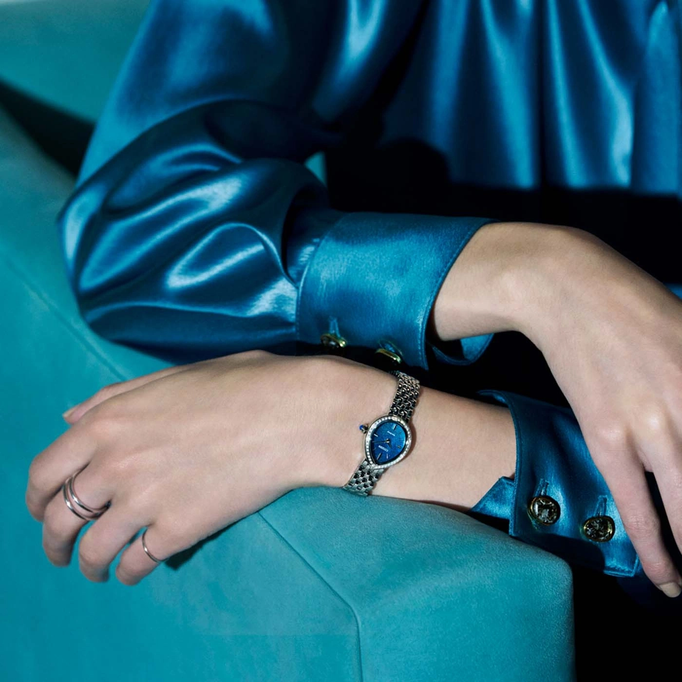 Eine Frau in einem blauen Satin-Oberteil und Ringen trägt eine Uhr.