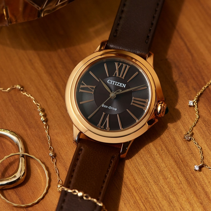 Citizen-Uhr mit braunem Lederarmband und goldenen Akzenten auf Holzoberfläche mit Schmuck.