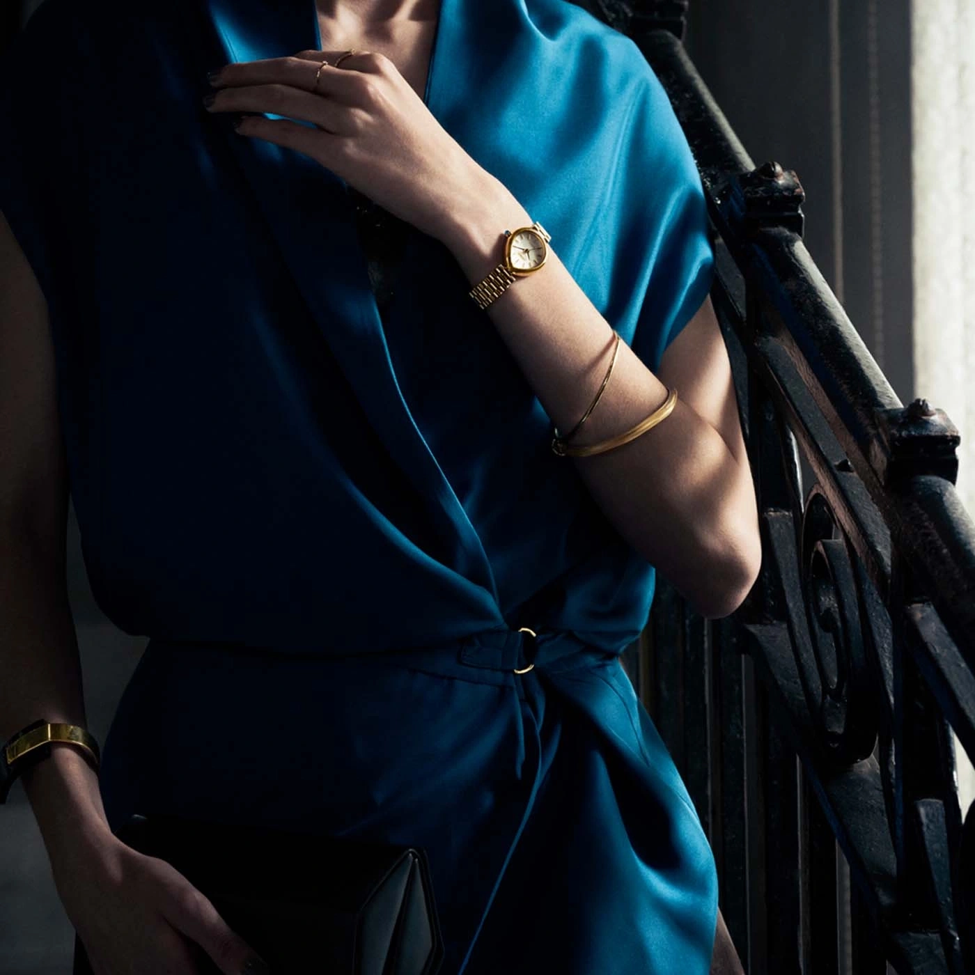 Frau im blauen Kleid mit Goldschmuck und Clutch.