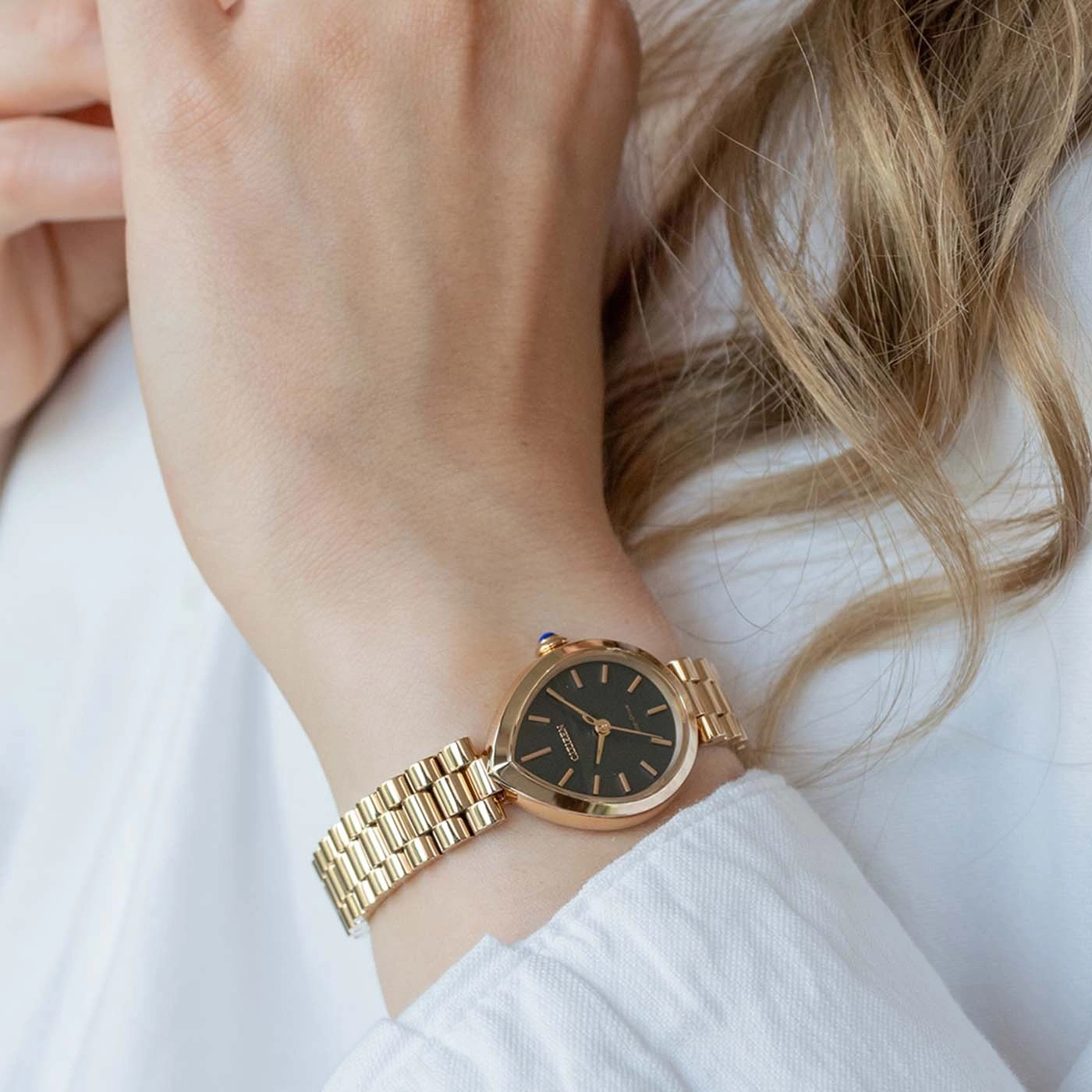 Goldene Uhr am Handgelenk einer Frau vor weißem Hemd.