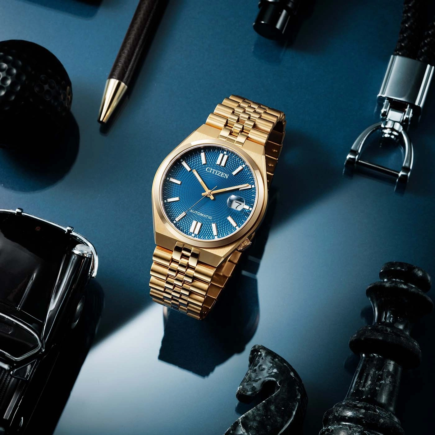 Goldene Uhr mit blauem Zifferblatt, umgeben von Accessoires auf blauem Untergrund.