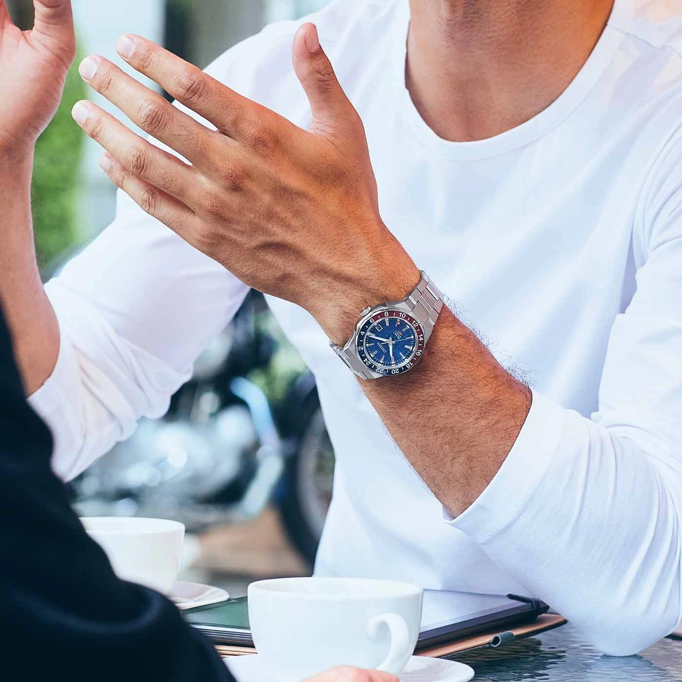 Mann mit Uhr, gestikuliert mit Händen, sitzt an einem Tisch mit Kaffeetassen.